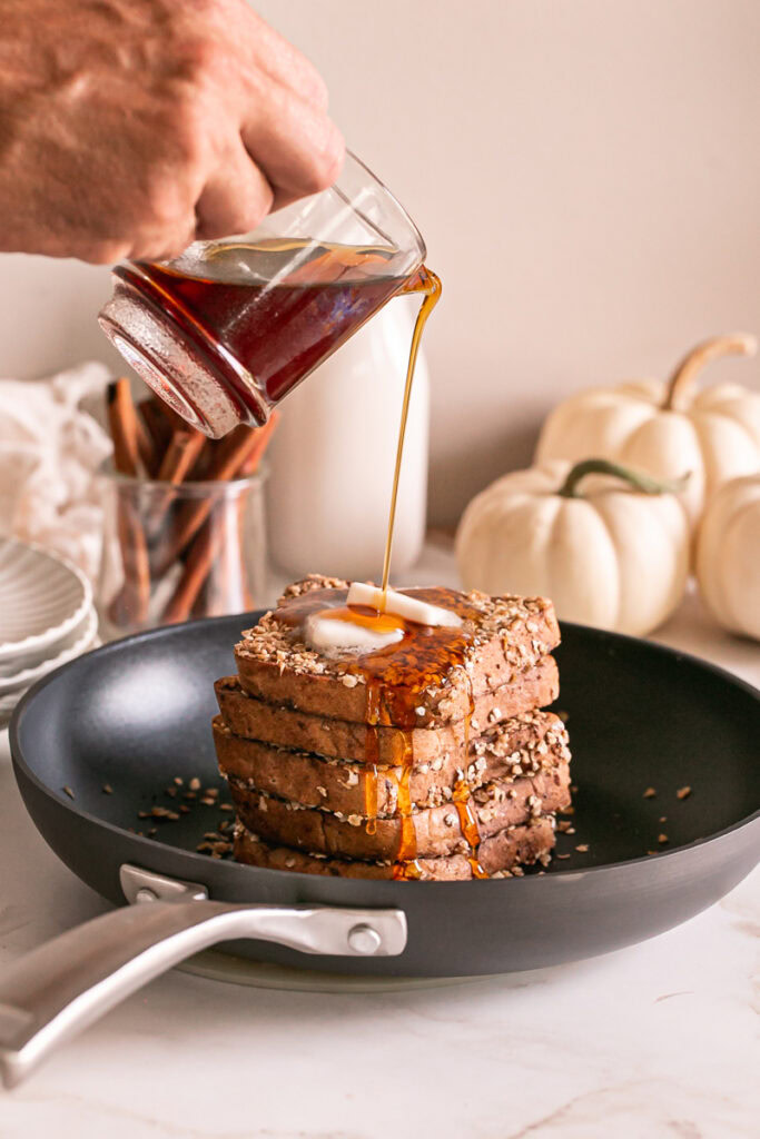 pumpkin spice oat crusted French toast stacked in a skillet with two squares of butter on the top one and drizzling maple syrup over the stack
