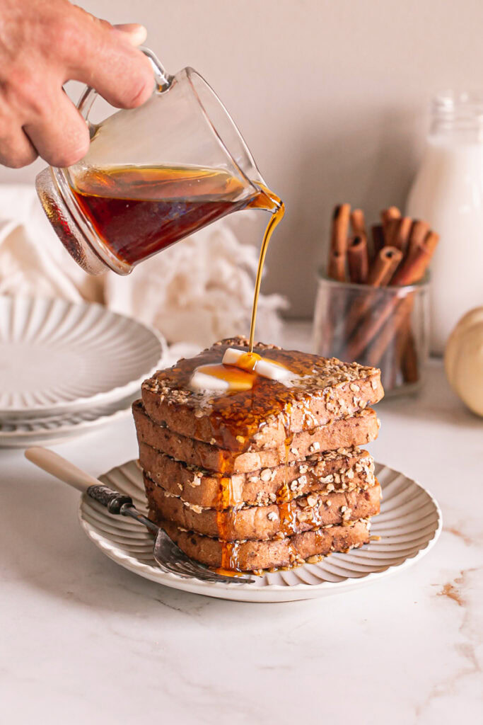 a stack of pumpkin spice oat French toast stacked on a plate with two squares of butter on the top slice. Maple syrup is being poured over top and running down the side