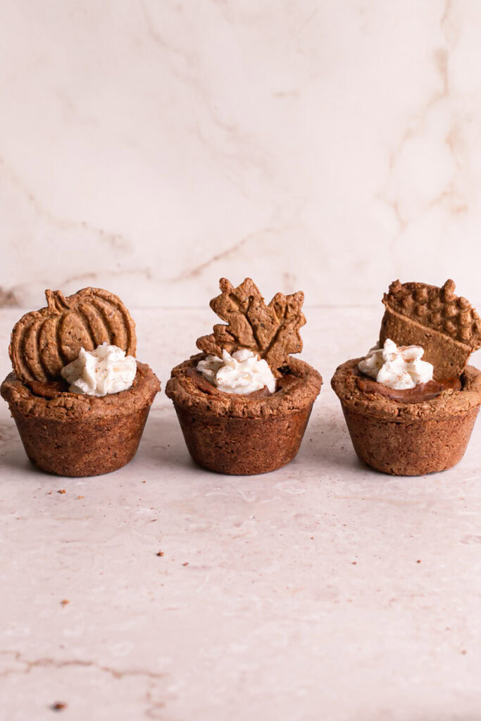 three pumpkin pie cookie cups in a row, one decorated with a pumpkin cookie, one with a maple leaf cookie, and the other with an acorn cookie